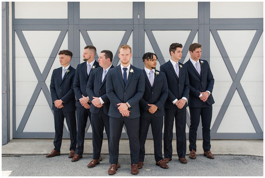 groom with his six groomsmen standing in a "V" by gray and white barn doors in navy suits at melhorn manor
