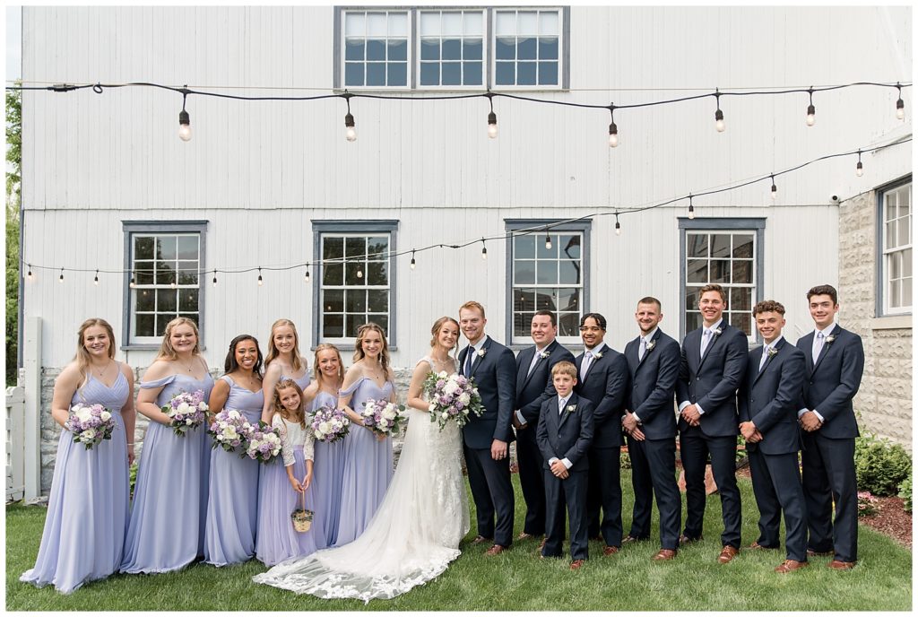 couple surrounded by their bridal party in front of white barn with outdoor string lights above them at melhorn manor in lancaster county