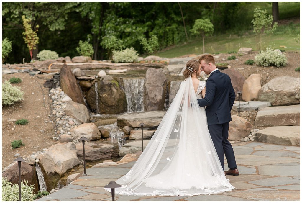 bride and groom with backs toward camera touching foreheads by landscaped rock fountain at melhorn manor