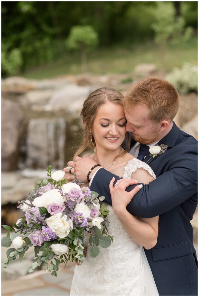 groom hugging bride from behind as she looks down holding bouquet by fountain in lancaster county