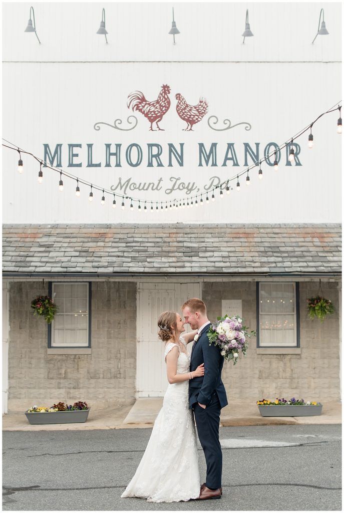 bride and groom hugging underneath string of lights and barn with "melhorn manor" painted on it in lancaster county pennsylvania