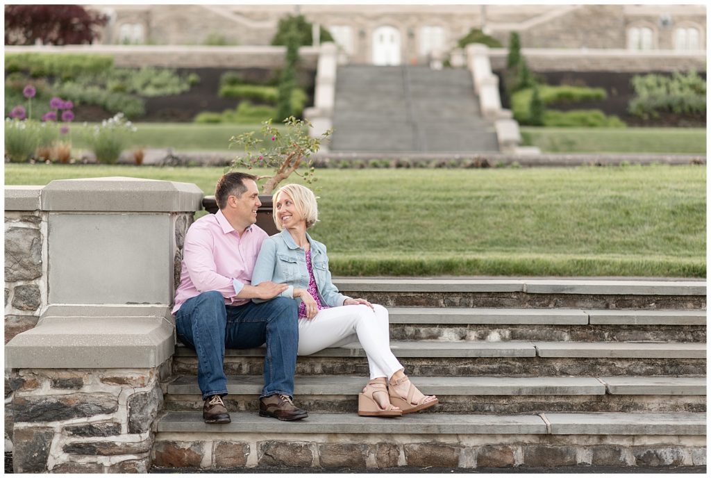 engaged couple sitting on beautiful stone steps with manicured lawn behind them as they smile at each other at masonic village