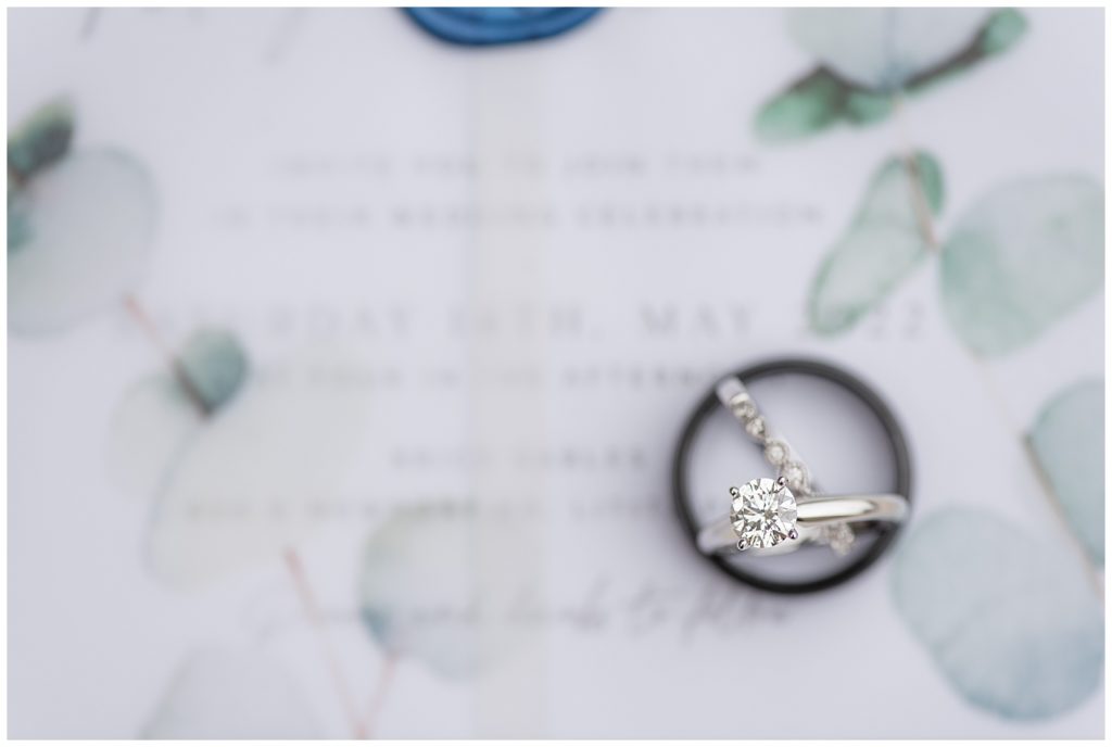 diamond wedding and engagement ring sitting inside black wedding band atop wedding invitation at brick gables
