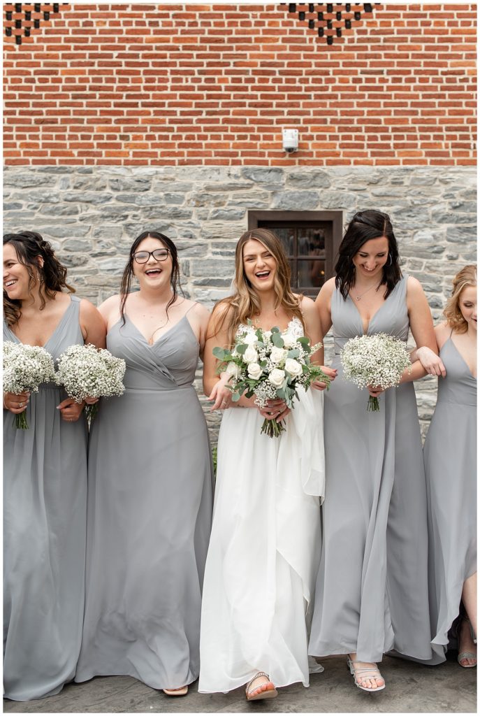 bride and bridesmaids walking with arms linked toward camera outside barn as they all laugh at brick gables in lancaster county