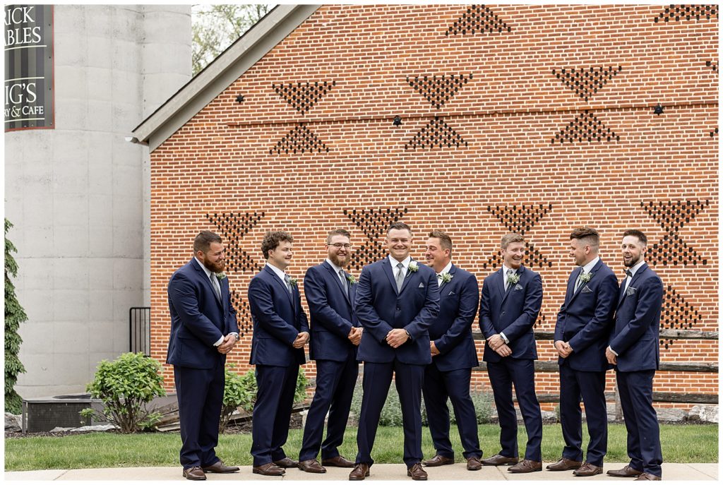 groom surrounded by his seven groomsmen all in navy blue suits outside brick barn in lititiz pennsylvania