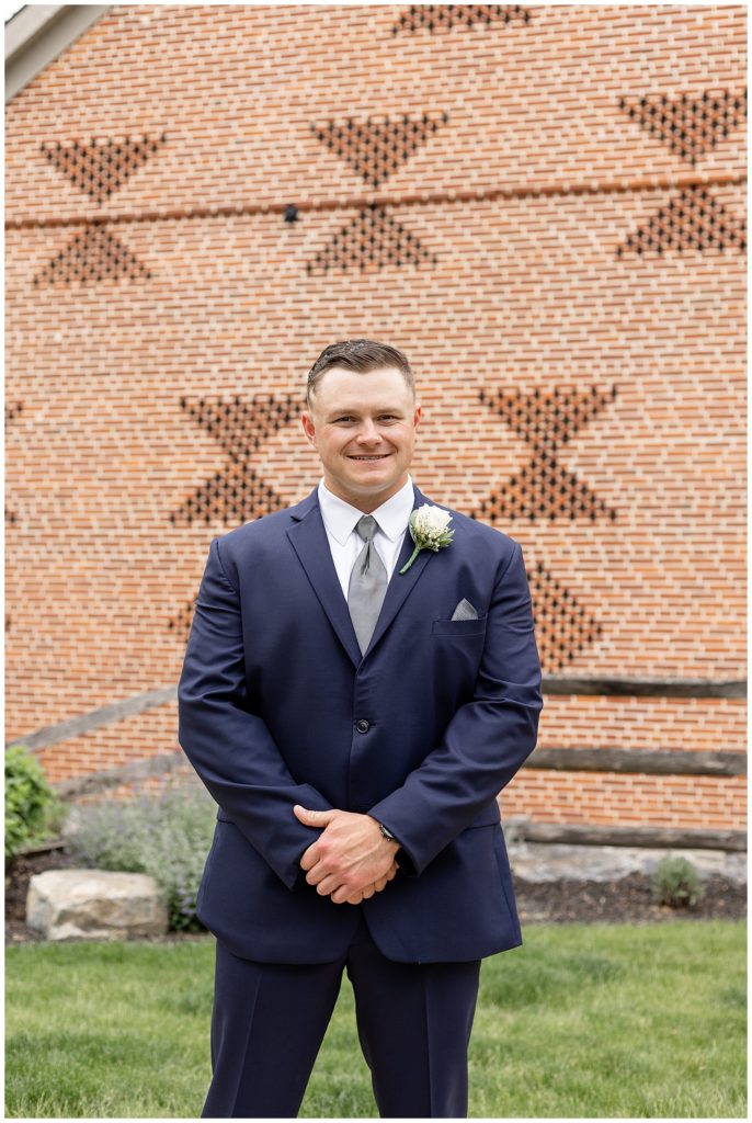 groom smiling at camera with his hands folded in front of him with brick barn in background in lititiz pennsylvania