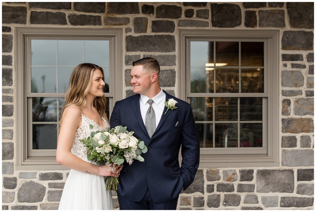 bride and groom smile as she holds large bouquet of white flowers outside barn in lancaster county