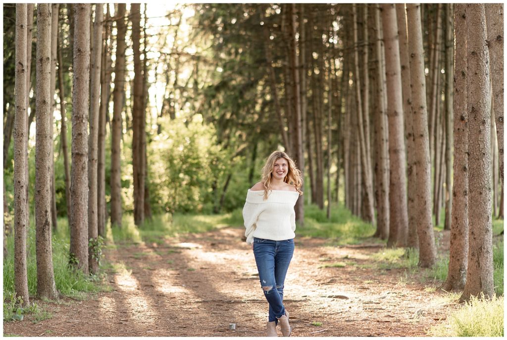 senior girl in white off-the-shoulder sweater and jeans standing between rows of pine trees at overlook park