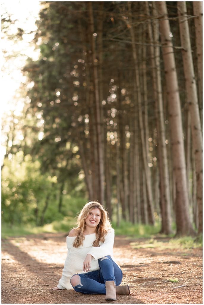 senior girl sitting on the ground by row of evergreen trees with left knee bent at overlook park