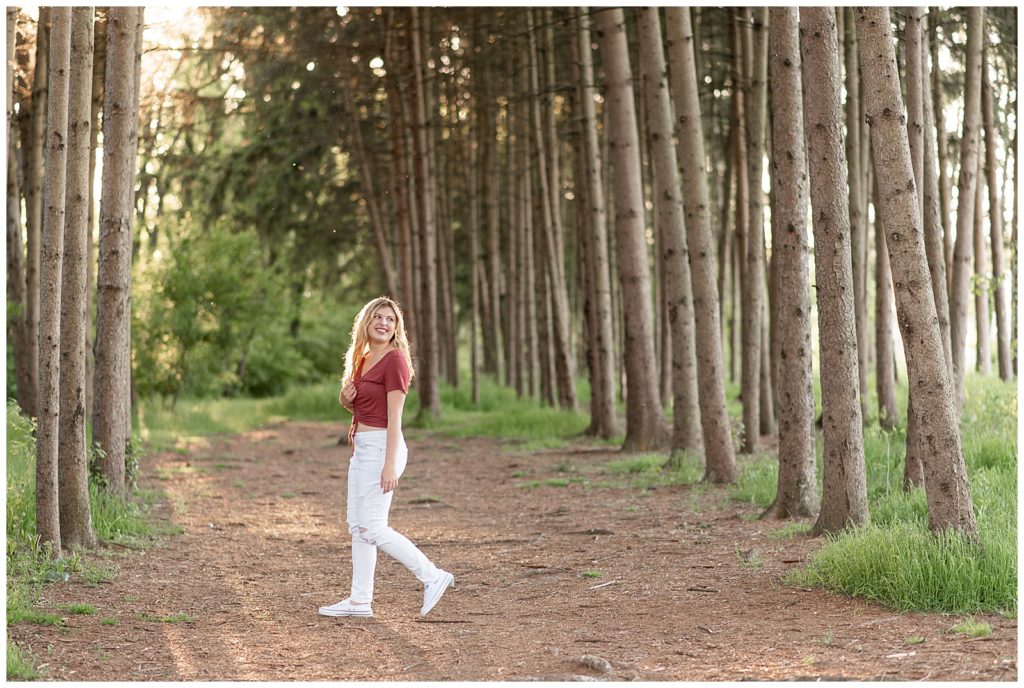 senior girl in rusty colored shirt with white jeans walking away from camera at sunset by row of trees at overlook park in lancaster pennsylvania