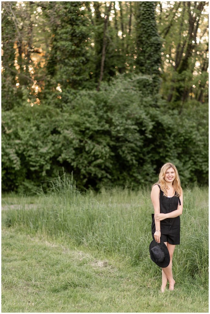 senior girl wearing little black dress and holding black trendy hat in her right hand smiling by tall grasses in manheim township