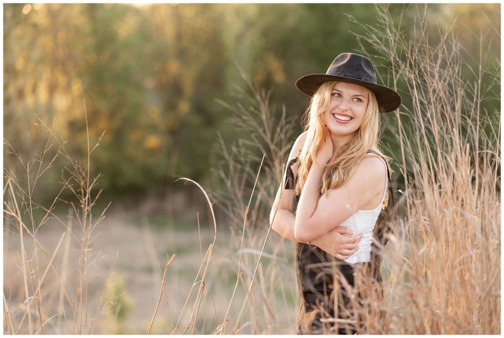 senior girl wearing trendy black hat with left hand on right shoulder and look back at sunset by tall grasses at overlook park