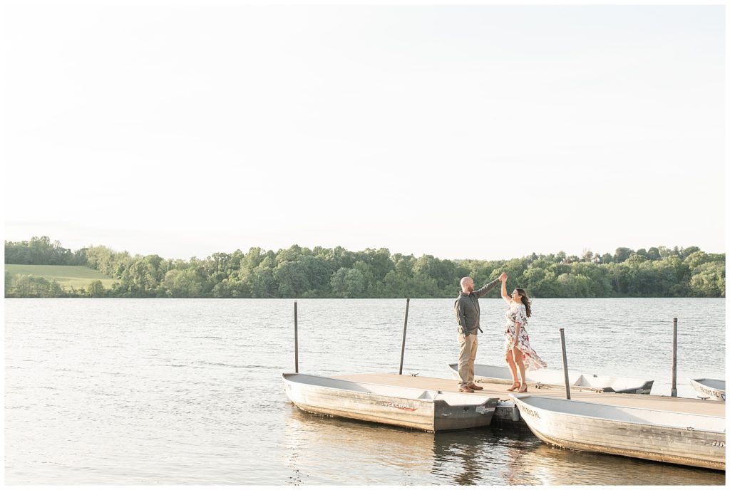 couple standing on dock by two boats as guy twirls girl under his left arm on sunny evening at marsh creek state park