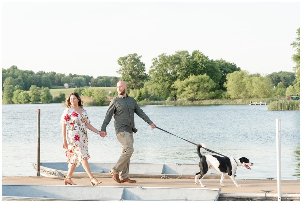 engaged couple holding hands and walking along boat dock with their dog leading the way at marsh creek state park