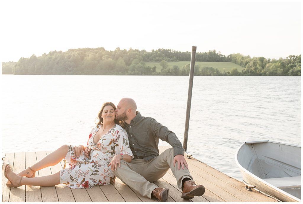 engaged couple sitting on the end of dock by water as guy kisses girl's left cheek on sunny evening at state park in chester county pennsylvania