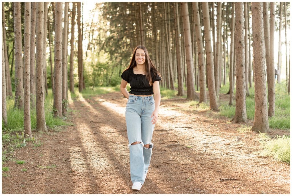 senior girl walking towards camera with right hand on hip by row of trees at overlook park