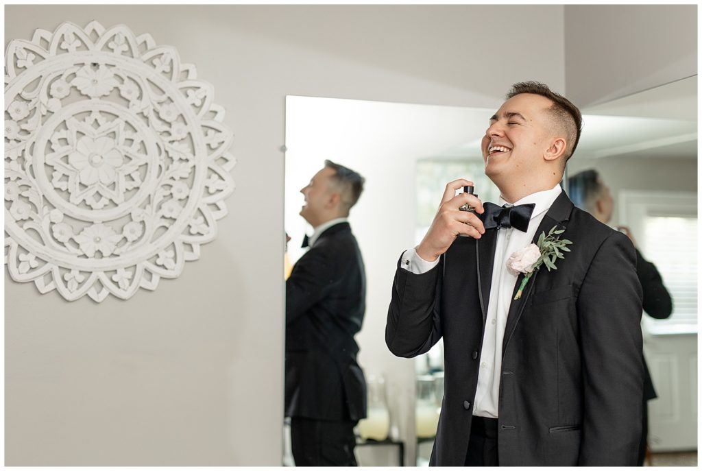 groom spritzing cologne onto his neck while wearing his black suit and bowtie in suite at cameron estate inn