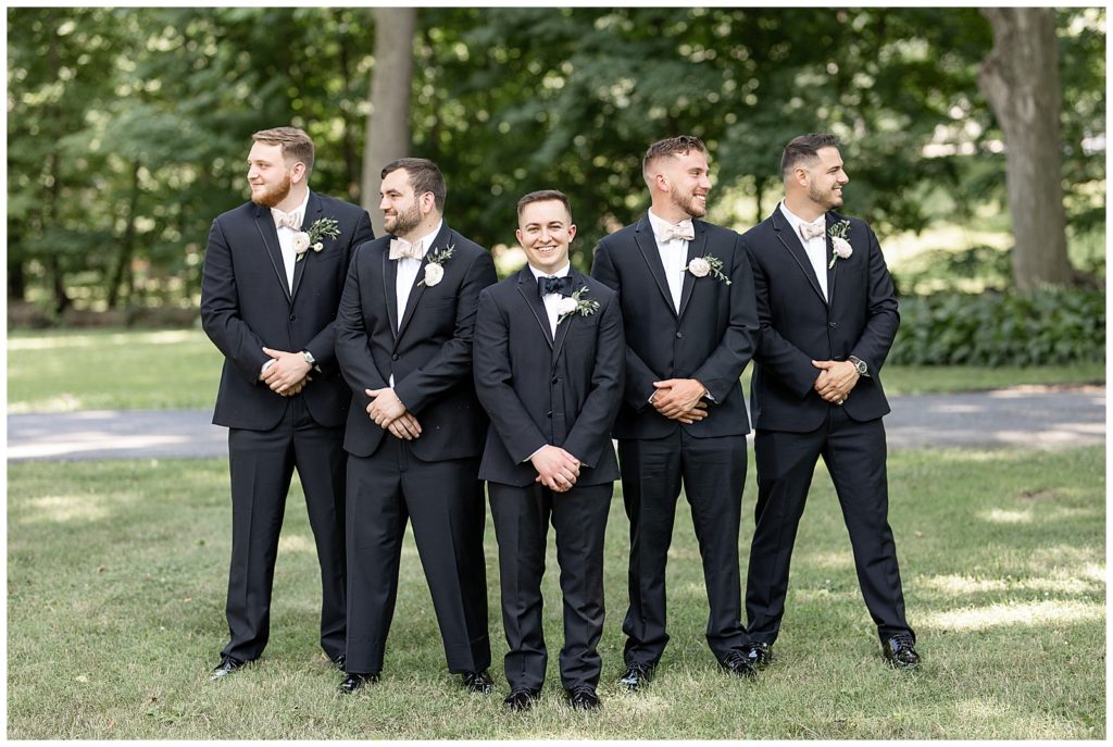 groom with his four groomsmen standing in a "v" behind him and looking in opposite directions under trees at cameron estate inn