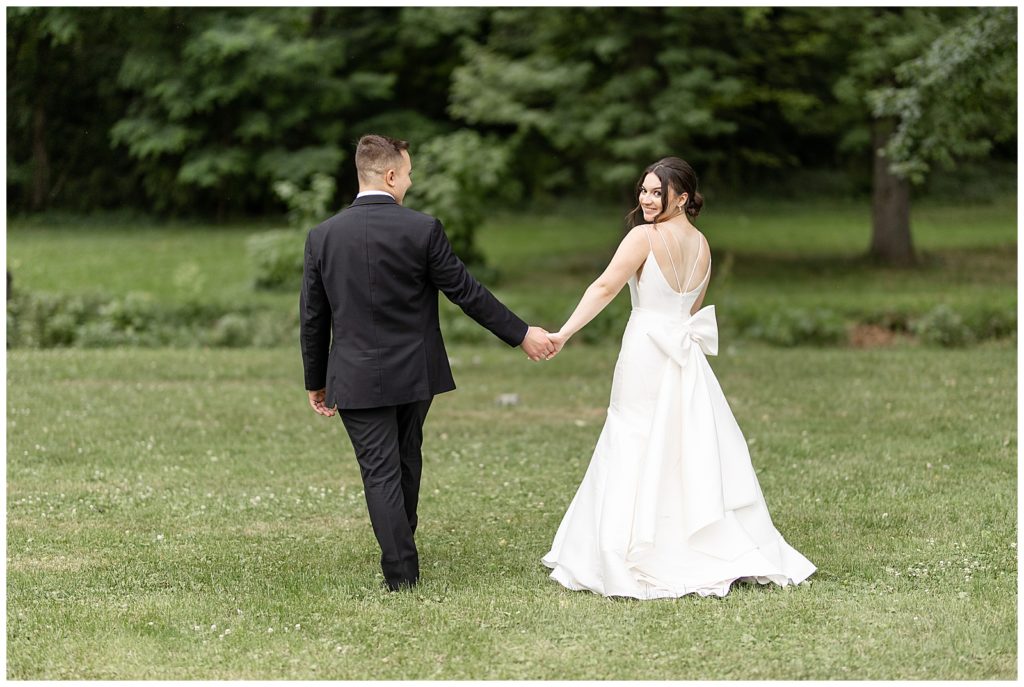 couple holding hands and walking away from camera as bride looks back over left shoulder on lawn at cameron estate inn