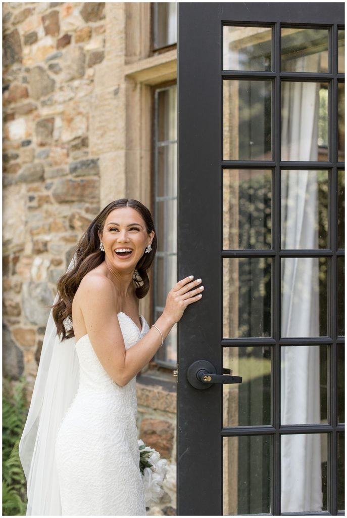 bride looking back and smiling big at camera as she holds black door open to stone building at parque ridley creek in pennsylvania