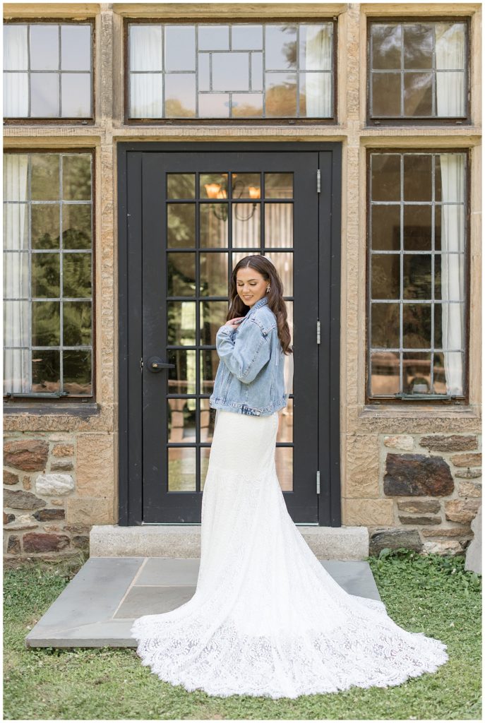 bride in her white wedding gown with blue jean jacket overtop as she looks down at left shoulder outside stone building at parque ridley creek wedding