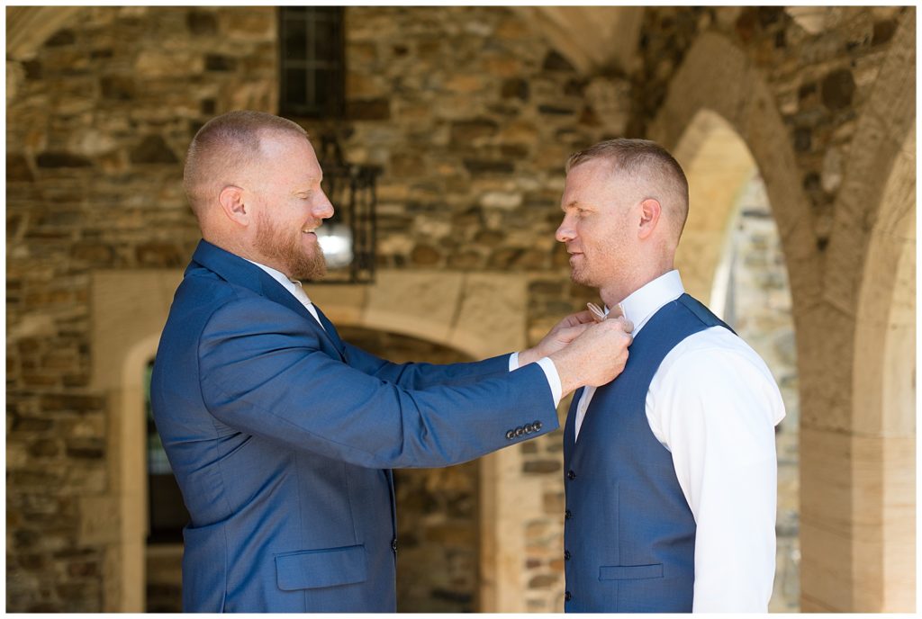best man adjusting groom's bowtie as they smile at each other wearing blue suits in delaware county pennsylvania