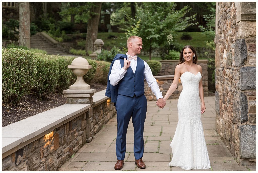 bride and groom holding hands as groom smiles at bride on stone walkway in garden in glen mills pennsylvania