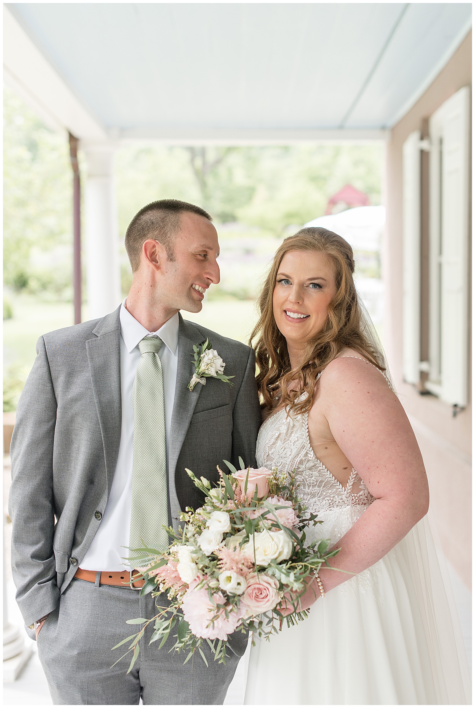 groom looking at his bride as she smiles at the camera holding her bouquet on front porch in lancaster county