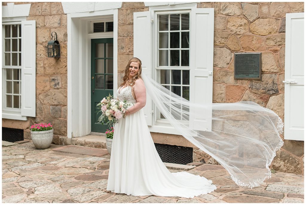 bride in beautiful white v-neck wedding gown with her long veil blowing behind her by home at elizabeth furnace
