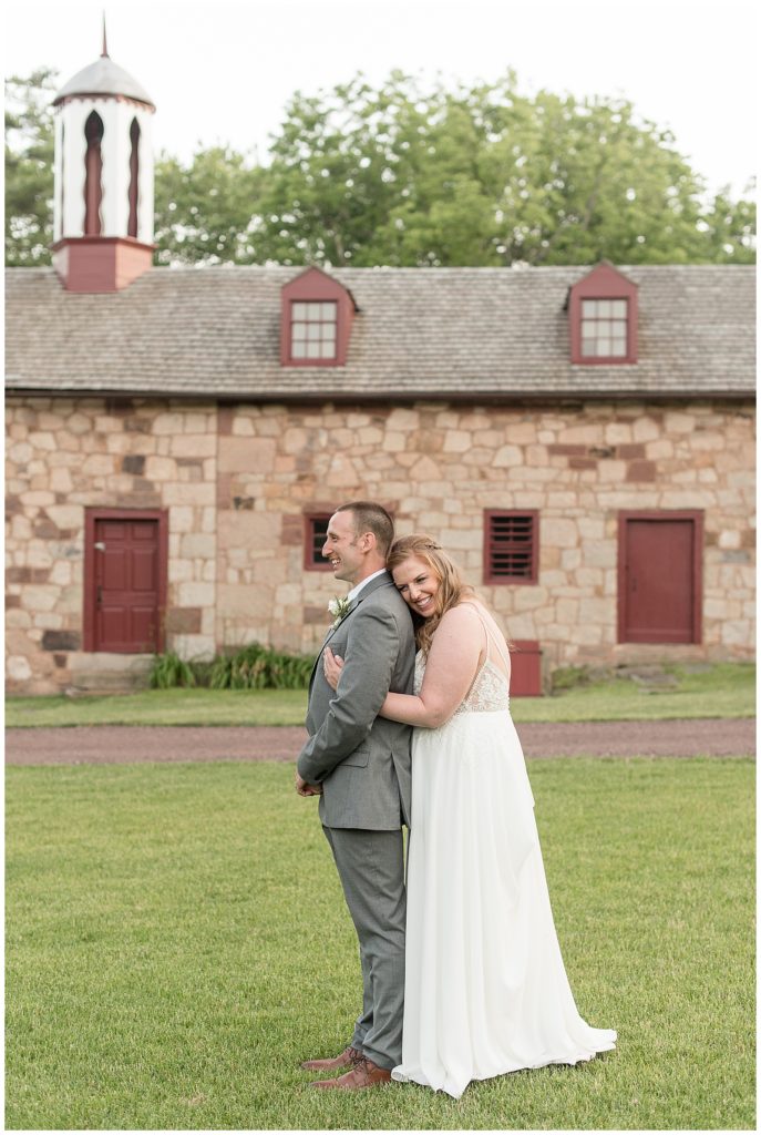 bride hugging her groom from behind as they both smile by stone barn at elizabeth furnace in lititz pennsylvania