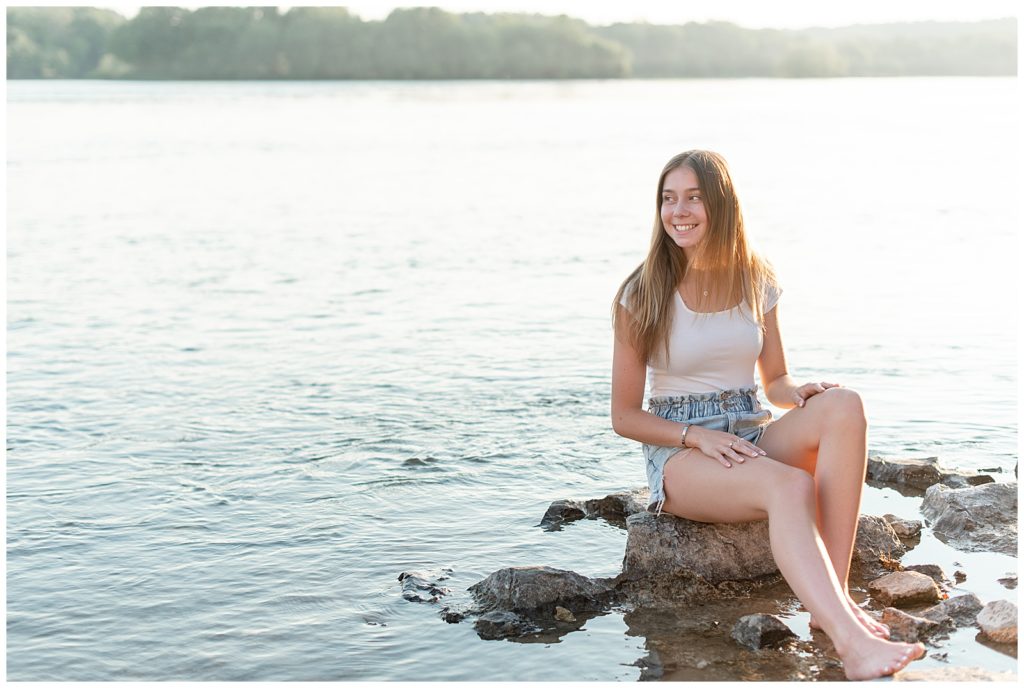 senior girl sitting on rock dipping her feet into shallow water of river as she looks right and smiles in lancaster county