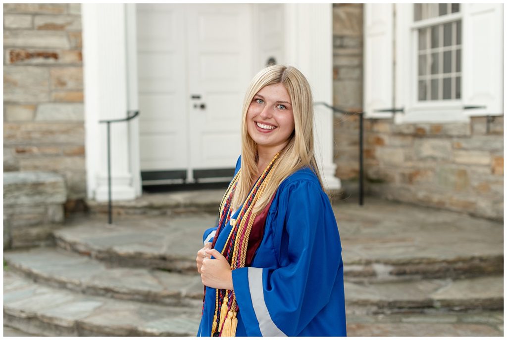 senior spokesmodel in blue graduation gown with left shoulder toward camera and smiling by stone building in lititz pennsylvania
