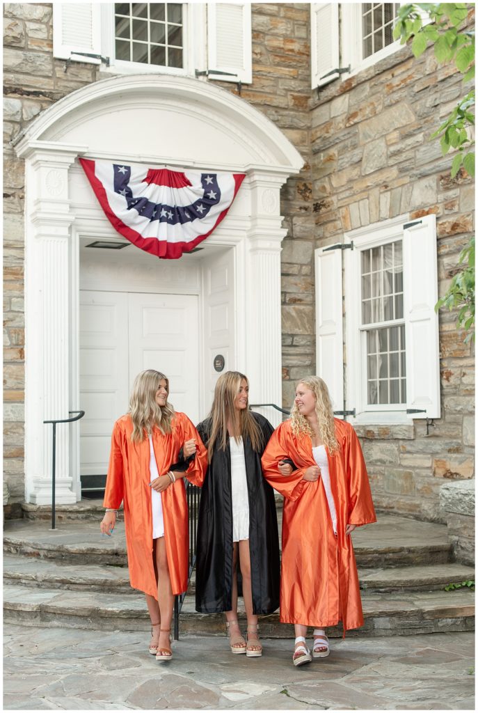 three class of 2022 senior girls in orange and black graduation gowns with arms linked by stone building in lititz pennsylvania