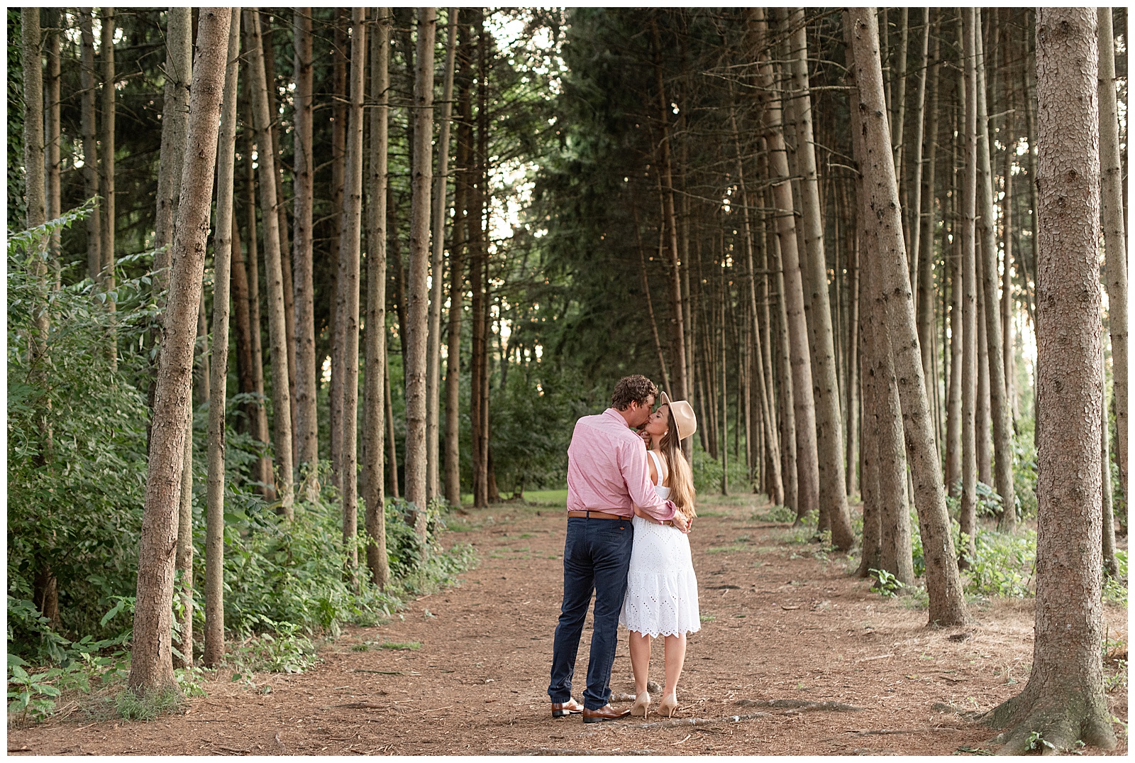 engaged couple with backs toward camera kissing by evergreen trees at manheim township park in lancaster county
