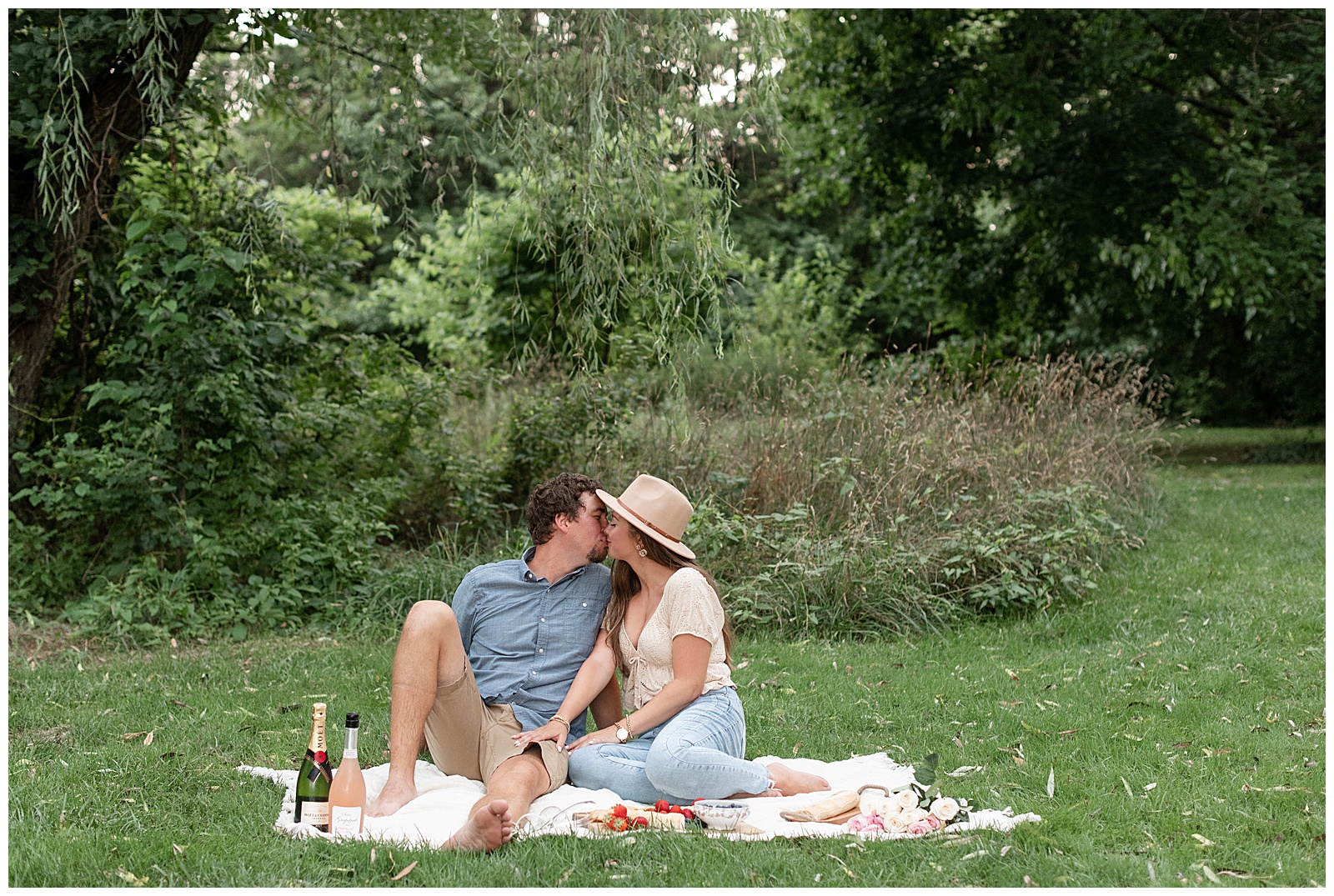 engaged couple kissing as they sit on blanket in the grass with champagne bottles in lancaster pennsylvania
