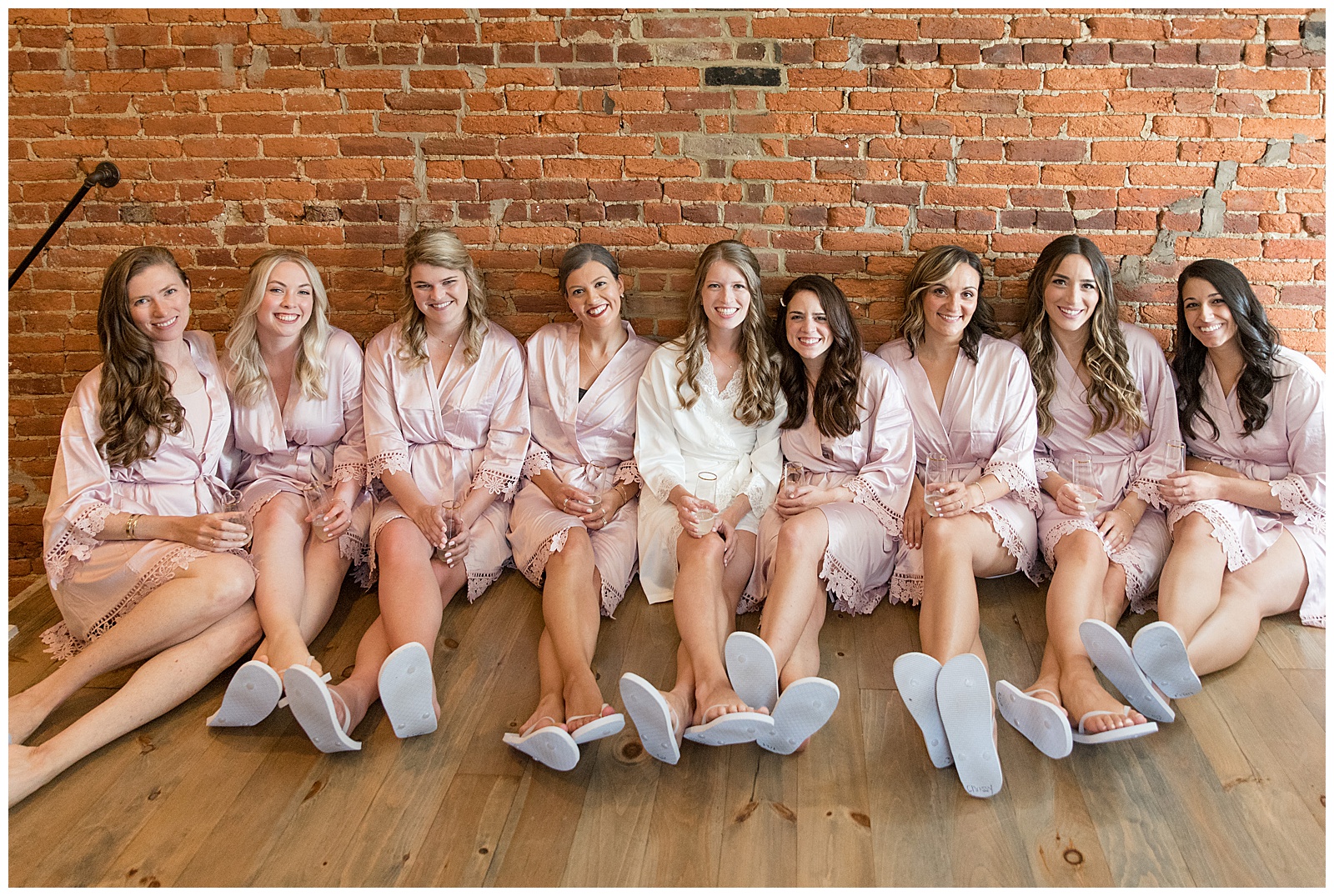 bride with her six bridesmaids sitting on the wood floor against a brick wall in their robes before the wedding at the barn at silverstone