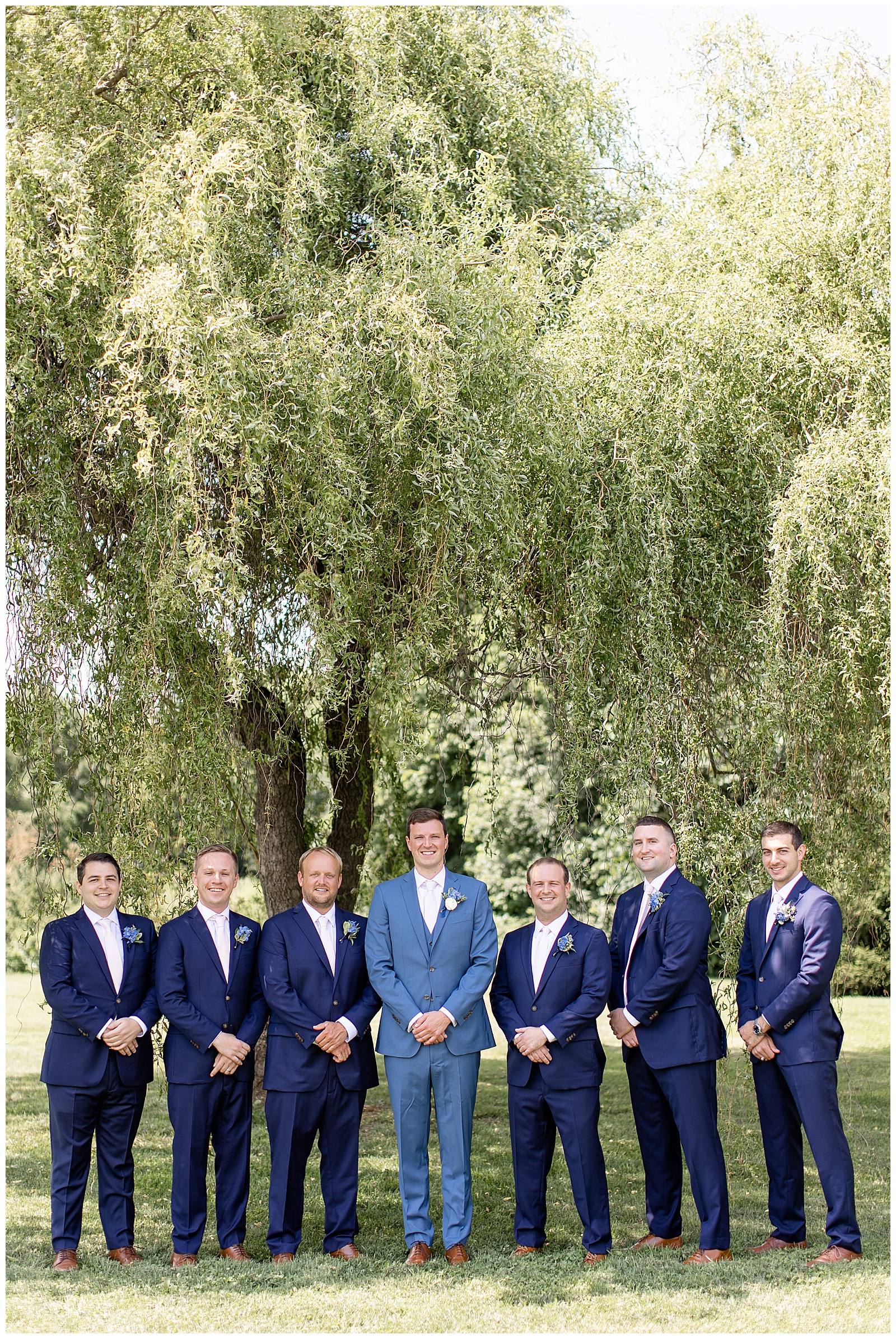groom with groomsmen under willow tree with their hands folded in front of them on sunny day in lancaster county