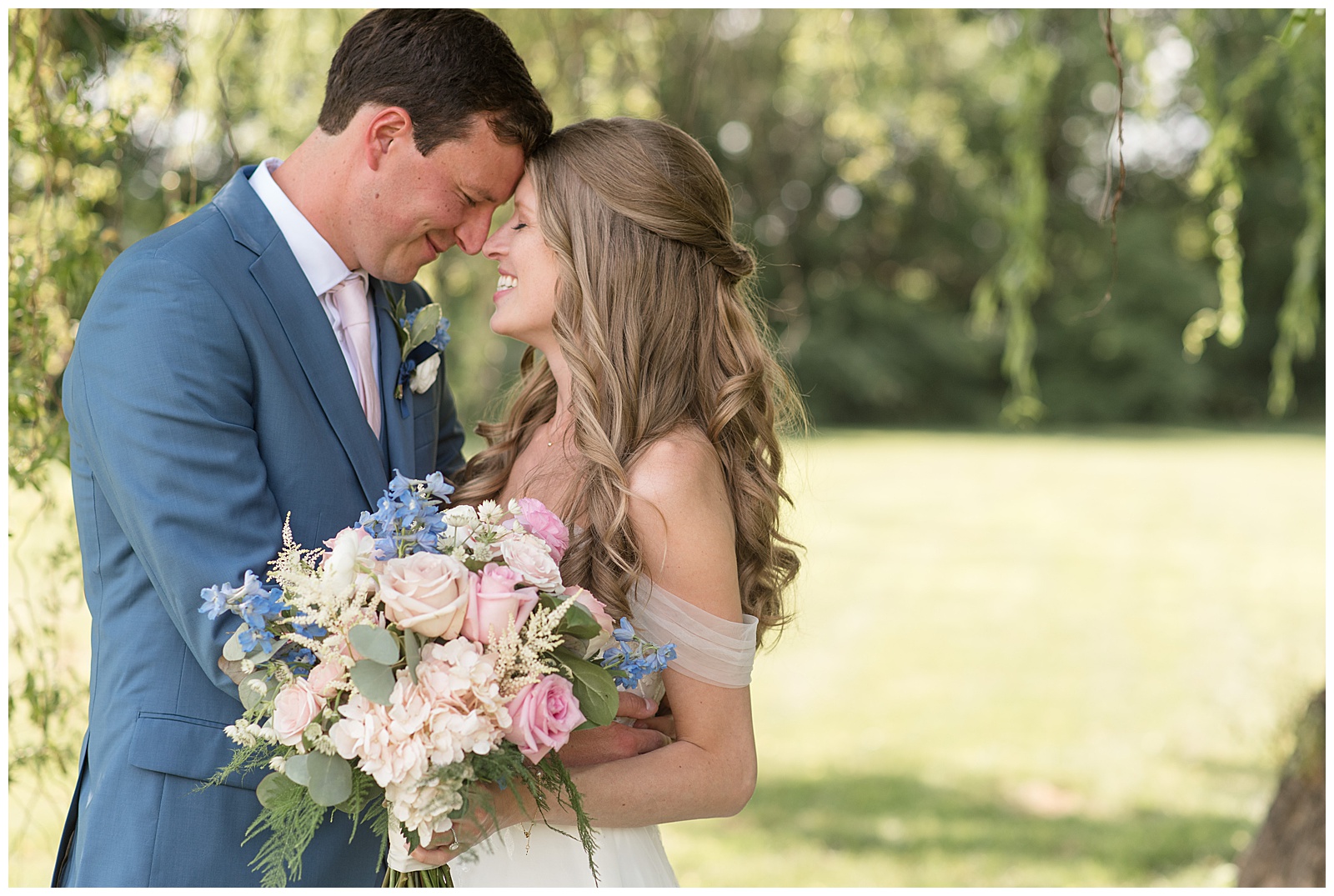 groom and bride resting their foreheads together as she holds bouquet by willow tree at the barn at silverstone