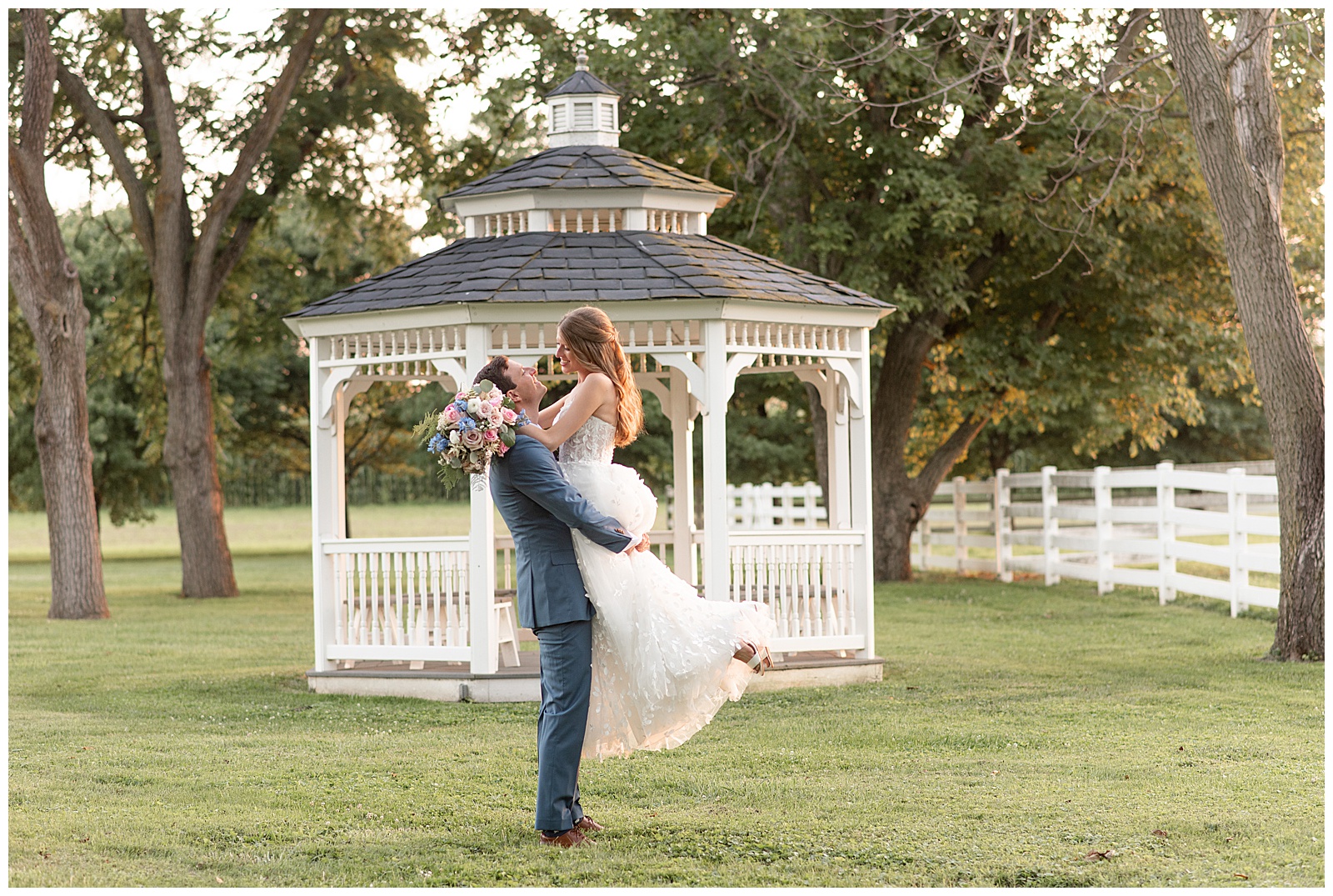 groom lifting his bride at sunset by white gazebo at the barn at silverstone