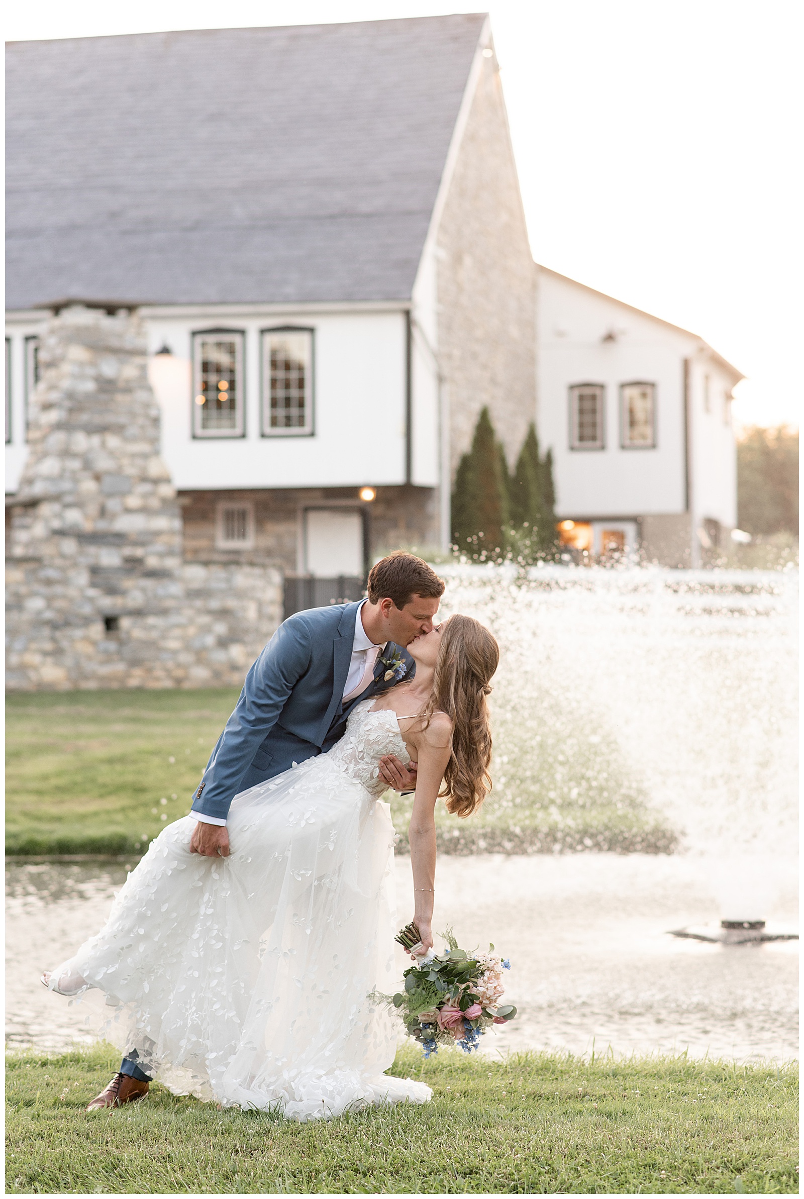 groom dipping his bride back and kissing her by pond and white barn at the barn at silverstone