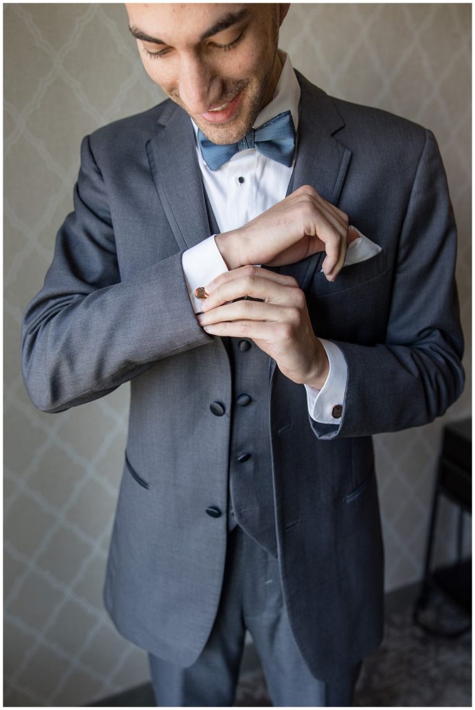 close up photo of groom adjusting his right cuff link while wearing dark gray suit and blue bow tie at normandy farms