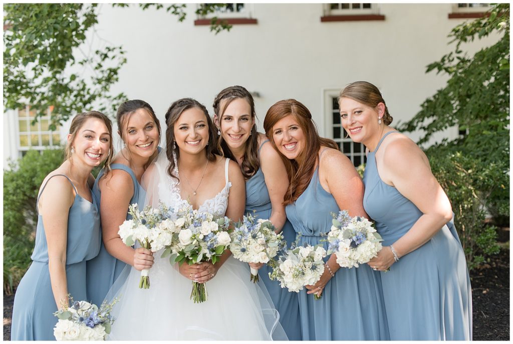 bride surrounded by her five bridesmaids wearing light blue gowns and all holding white and blue bouquets at normandy farms