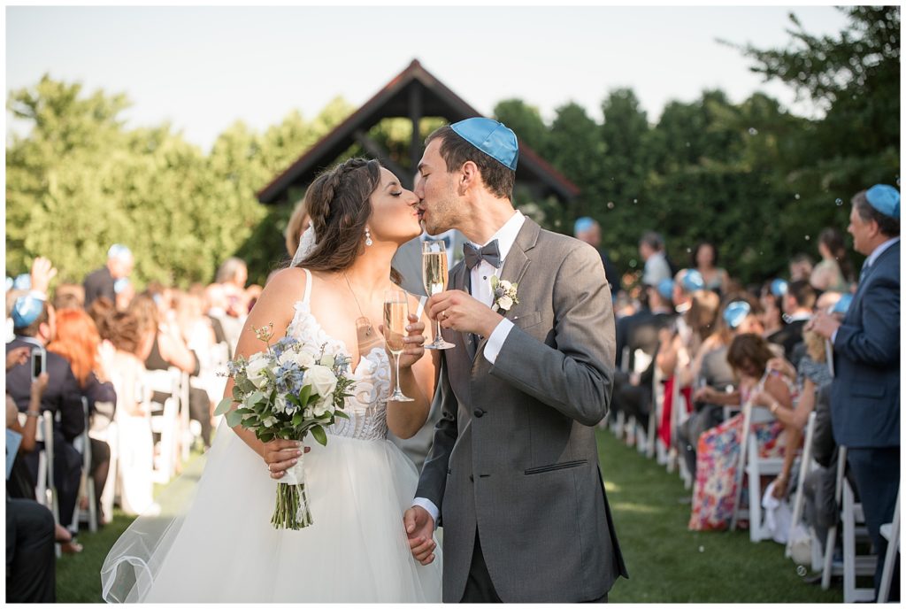 bride and groom kissing while holding glasses of champagne at the end of aisle at outdoor ceremony at normandy farms