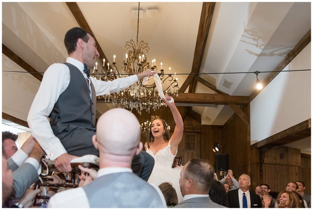 bride and groom sitting on chairs and being raised up by guests during jewish dance at reception at normandy farms