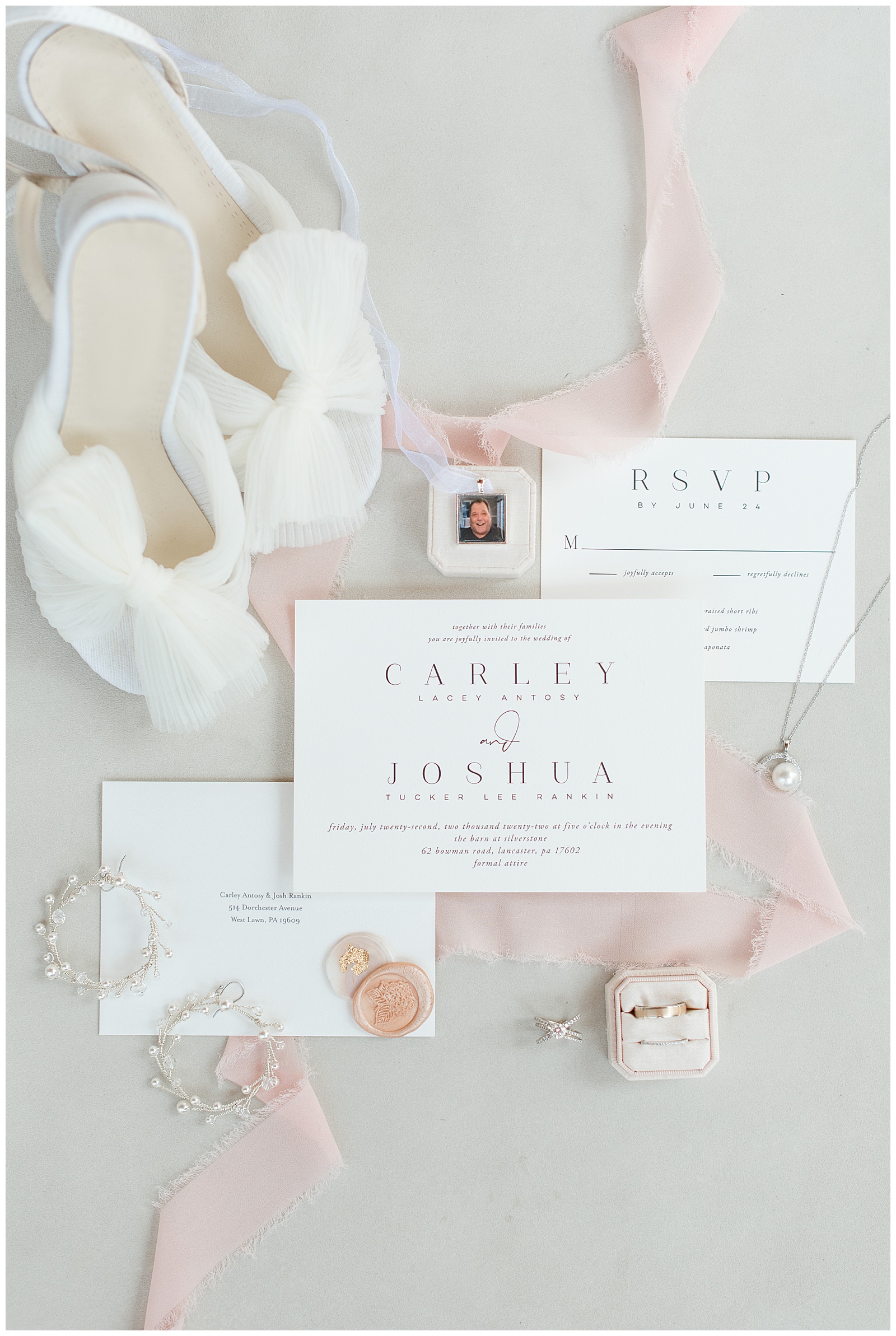 wedding invitation surrounded by light pink ribbon, bride's white sandals, wedding rings and jewelry at the barn at silverstone
