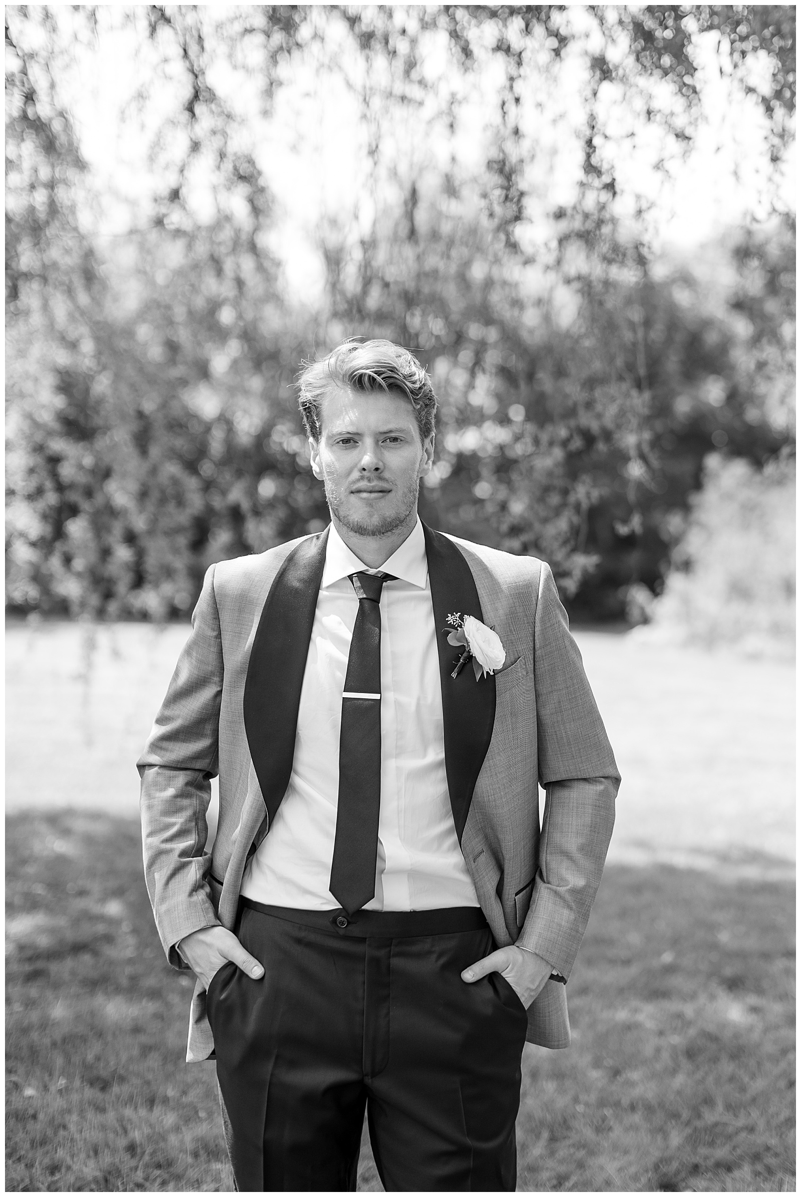 black and white photo of groom with his hands in his pockets by willow tree at the barn at silverstone