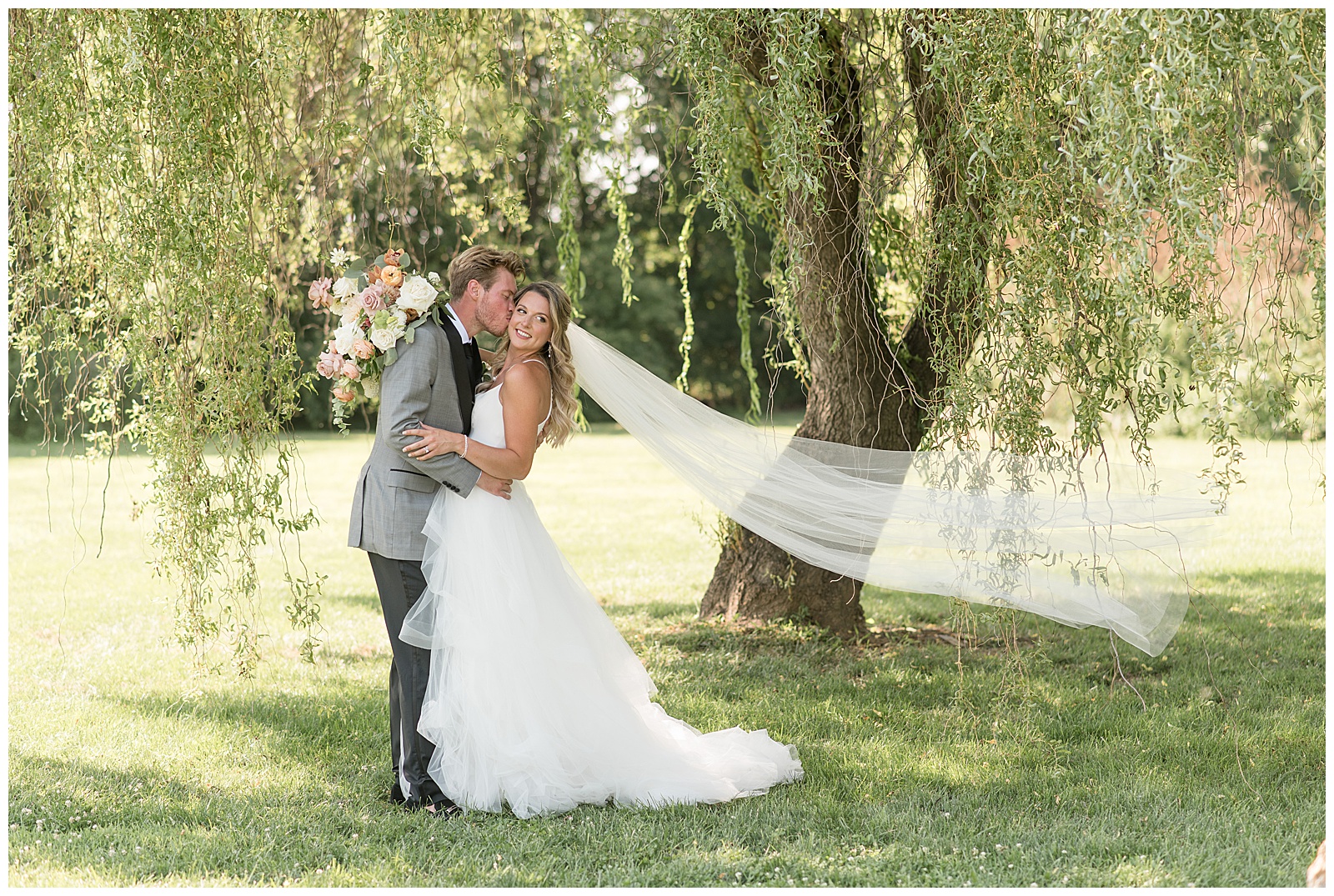 groom kissing his bride on her right cheek as she smiles with long veil blowing behind her under willow tree in lancaster county