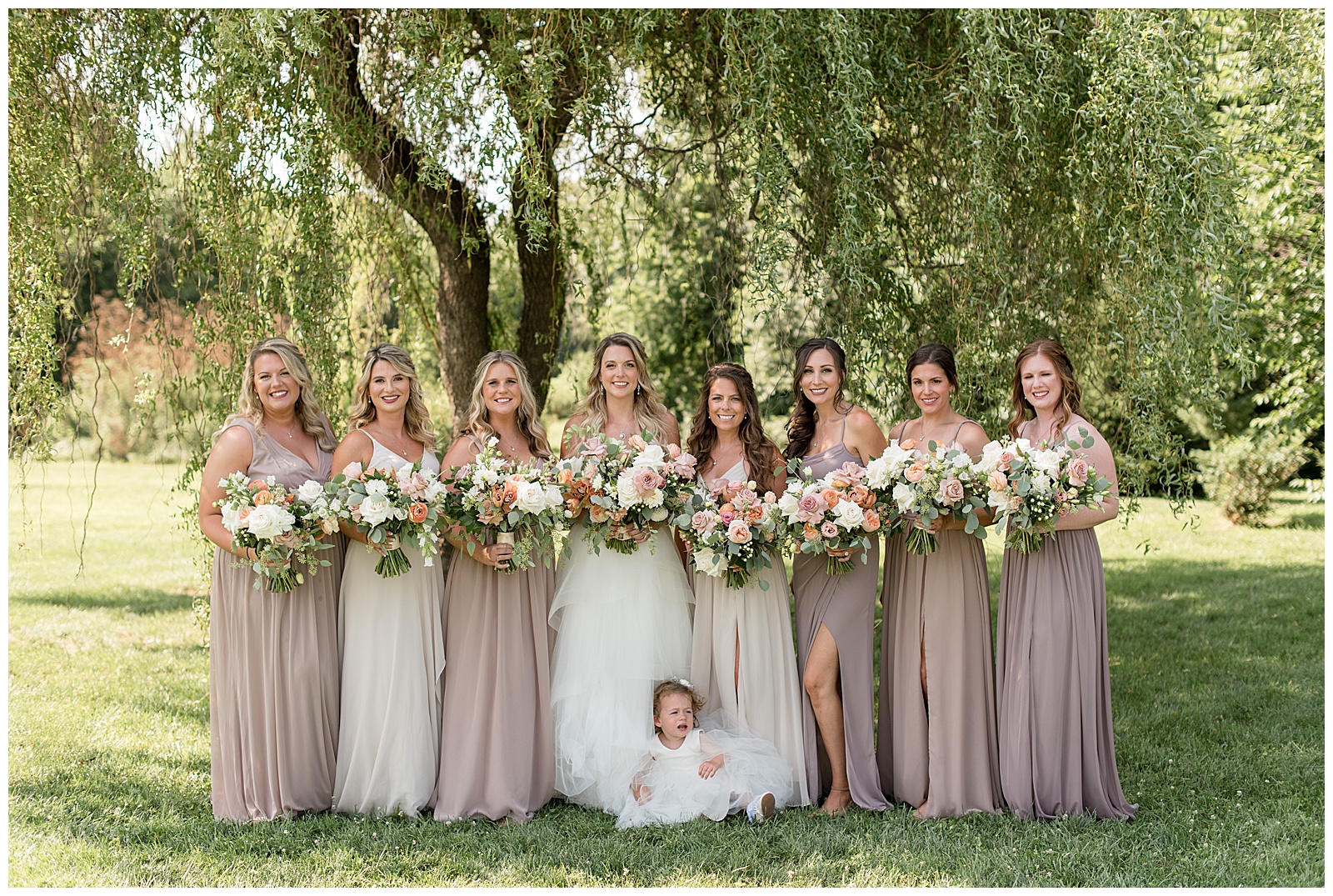 bride with her seven bridesmaids and tiny flower girl in mauve and white gowns under willow tree at the barn at silverstone
