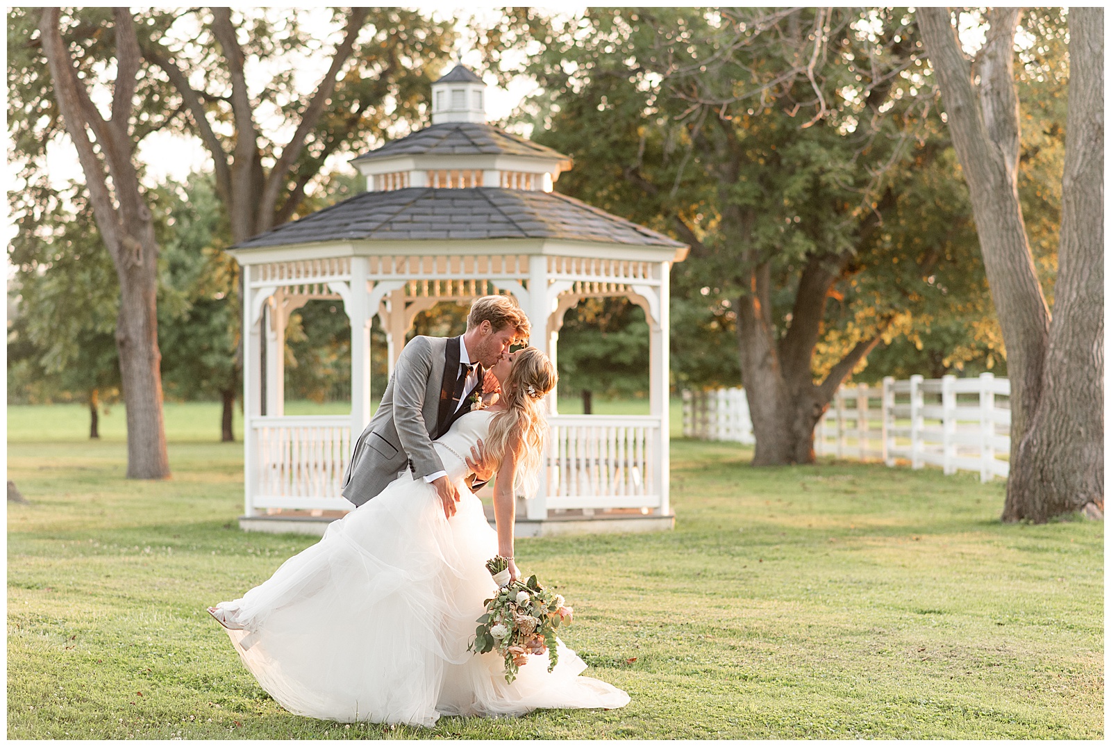 groom dips his bride back as they kiss by white gazebo at sunset at the barn at silverstone