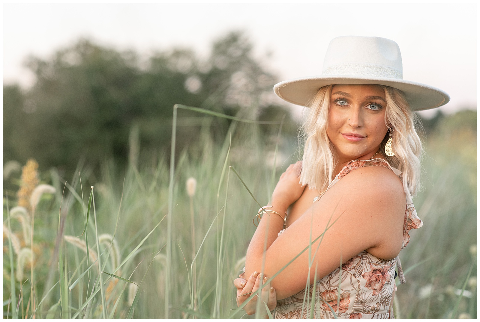 senior girl wearing floral dress and white brimmed hat with left hand on right shoulder in field of tall grasses at sunset in lancaster pa